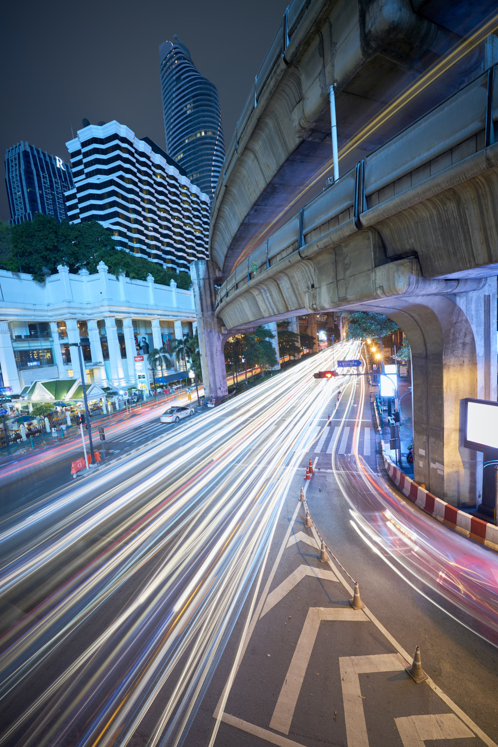 Bangkok, Thailand: Creative high angle night shot of Ratchaprasong Intersection and Erawan Shrine, with colourful light trail from cars driving past in busy traffic.